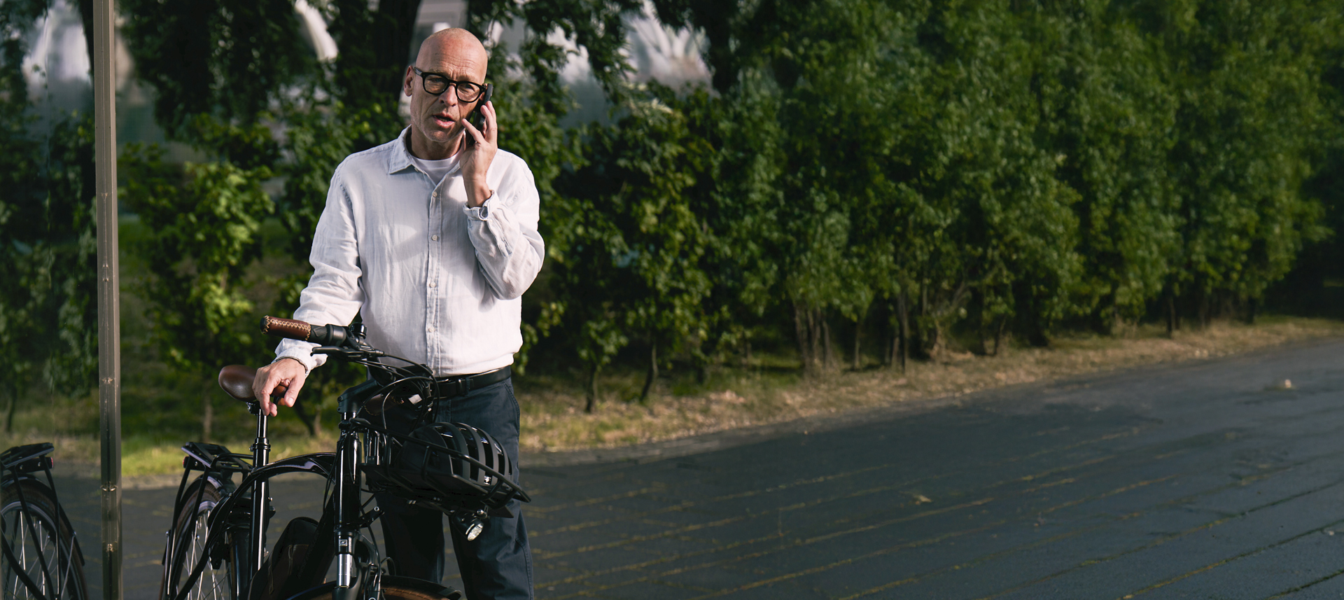 Ein Mann steht neben einem Fahrrad am Straßenrand und telefoniert mit seinem Smartphone. Im Hintergrund sind Bäume und eine ruhige Straße zu sehen.