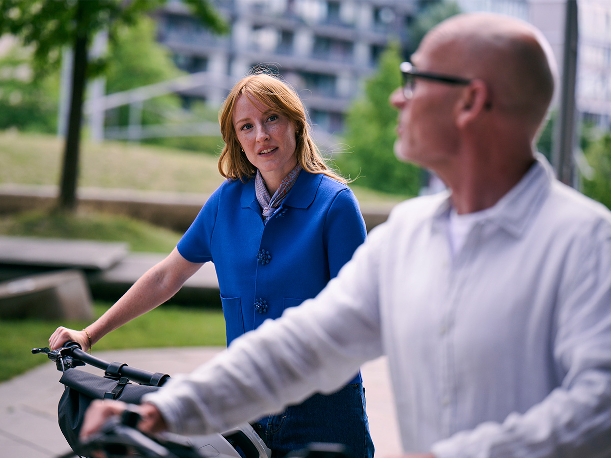 Eine Frau und ein Mann stehen im Freien neben ihren Fahrrädern und unterhalten sich. Im Hintergrund sind Grünflächen und Gebäude zu sehen.