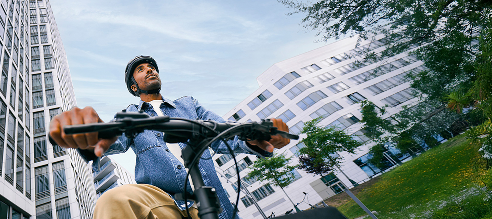 Ein Mann mit Fahrradhelm fährt mit einem Fahrrad durch eine moderne Stadtumgebung. Die Aufnahme ist aus niedriger Perspektive und zeigt im Hintergrund hohe Gebäude und Grünflächen.