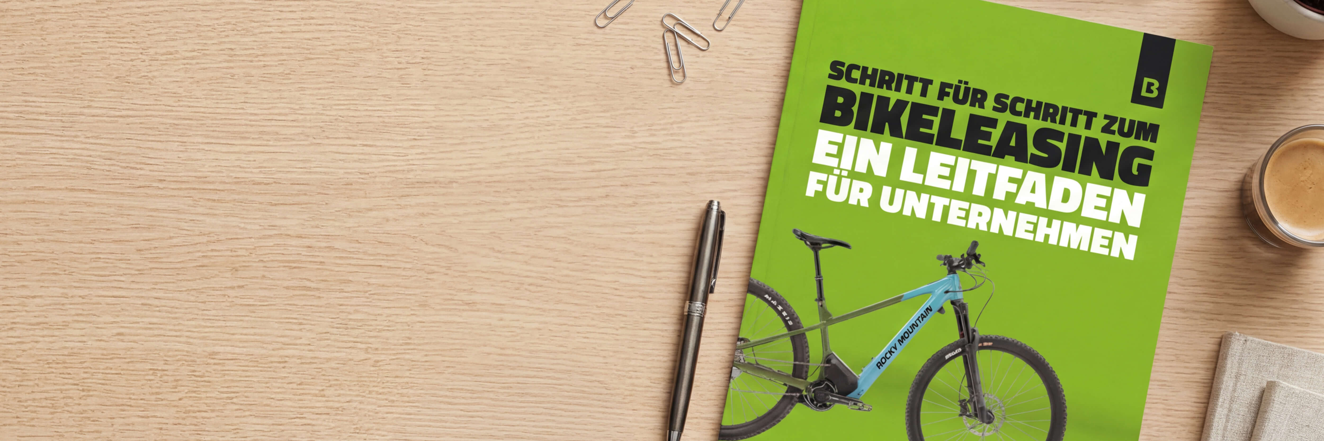 Ein Leitfaden zum Bikeleasing für Unternehmen liegt auf einem Holztisch neben einem Stift, Büroklammern und einer Tasse Kaffee.