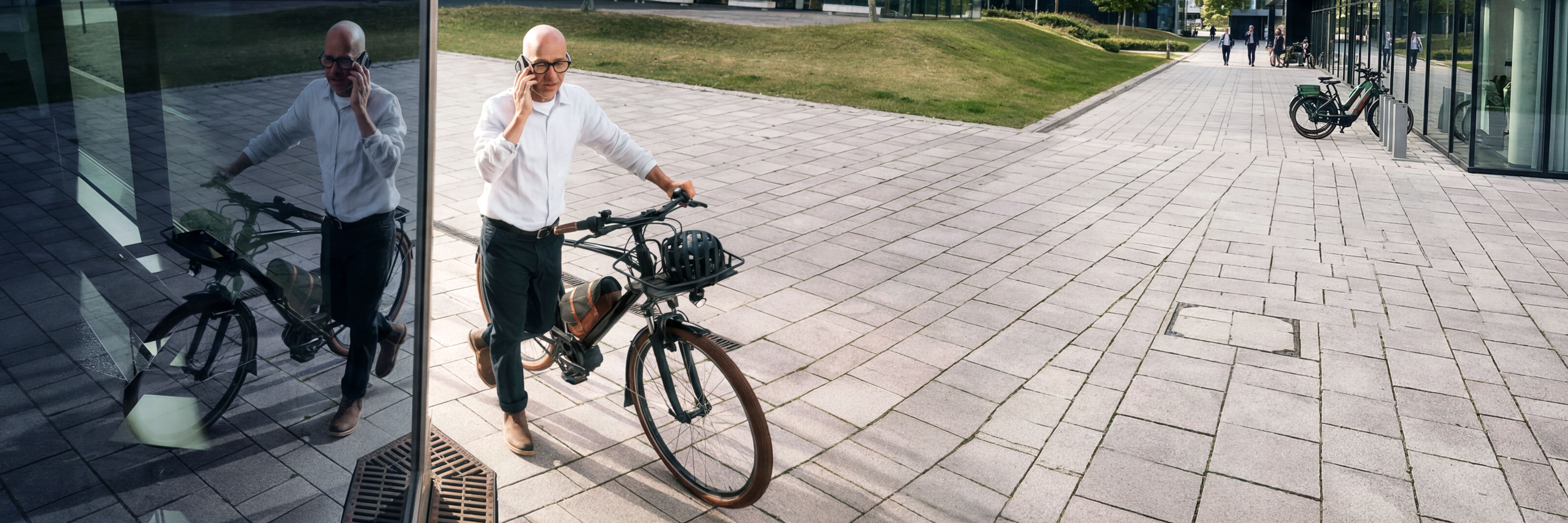 Ein Mann schiebt ein Fahrrad über einen gepflasterten Weg und telefoniert dabei mit seinem Smartphone. Die Szene spiegelt sich in einer Glasfassade, im Hintergrund sind Gebäude und weitere Fahrräder zu sehen.
