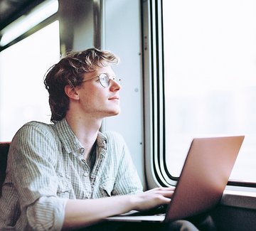 Mann sitzt mit einem Laptop in der Bahn und schaut aus dem Fenster