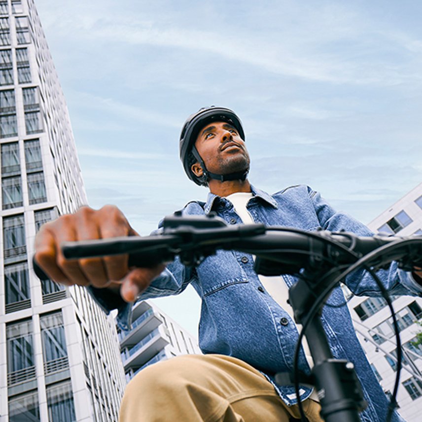 Ein Mann mit Fahrradhelm fährt mit einem Fahrrad durch eine moderne Stadtumgebung. Die Aufnahme ist aus niedriger Perspektive und zeigt im Hintergrund hohe Gebäude und Grünflächen.