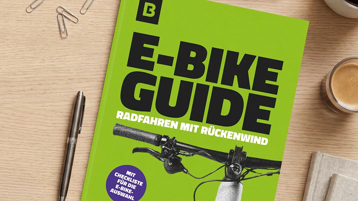 Auf einem Holztisch liegt ein grünes Whitepaper mit dem Titel „E‑Bike Guide – Radfahren mit Rückenwind“ und einer Lenker-Abbildung; daneben sind ein Stift, Büroklammern und eine Kaffeetasse zu sehen.