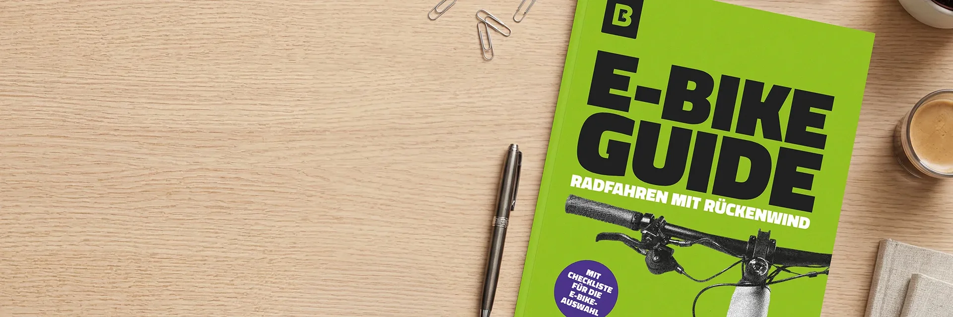 Auf einem Holztisch liegt ein grünes Whitepaper mit dem Titel „E‑Bike Guide – Radfahren mit Rückenwind“ und einer Lenker-Abbildung; daneben sind ein Stift, Büroklammern und eine Kaffeetasse zu sehen.