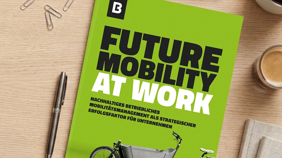 Auf einem Holztisch liegt ein grünes Whitepaper mit dem Titel „Future Mobility at Work“; daneben sind ein Stift, Büroklammern und eine Kaffeetasse zu sehen.