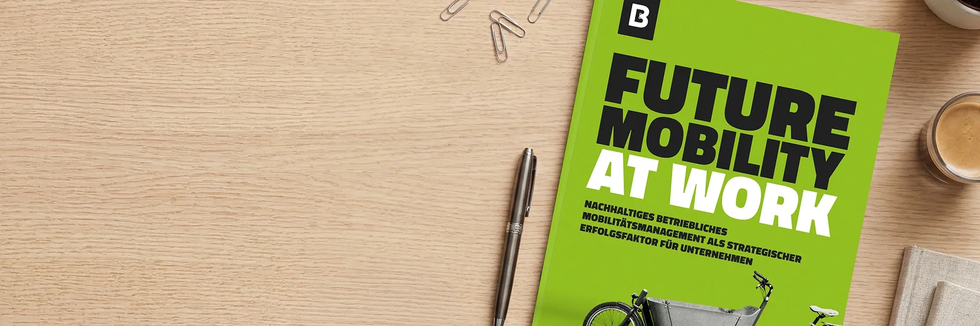 Auf einem Holztisch liegt ein grünes Whitepaper mit dem Titel „Future Mobility at Work“; daneben sind ein Stift, Büroklammern und eine Kaffeetasse zu sehen.