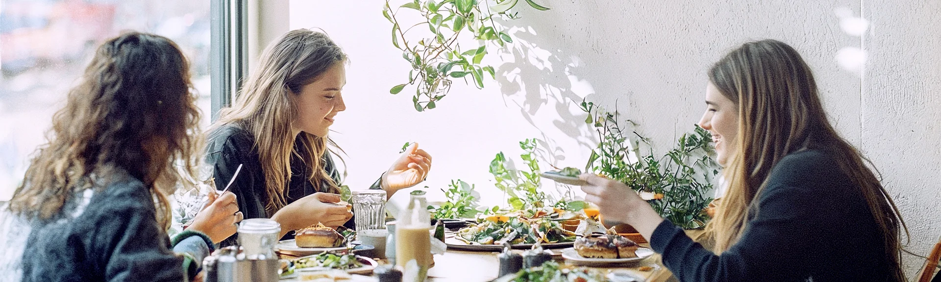 Drei Frauen sitzen an einem Tisch in einem hellen Café oder Pausenraum und essen gemeinsam zu Mittag. Auf dem Tisch stehen mehrere Teller mit Speisen und Getränke, im Hintergrund hängen Pflanzen an der Wand und ein großes Fenster sorgt für Tageslicht.