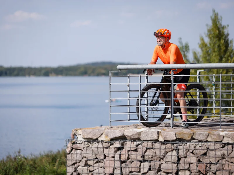 Ein Radfahrer in orangefarbener Sportkleidung steht mit seinem Fahrrad an einer Aussichtsplattform am Wasser. Im Hintergrund sind ein See und Bäume zu sehen.