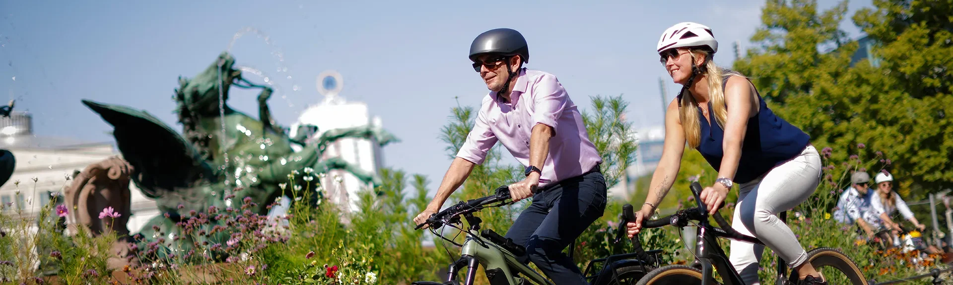 Zwei Personen fahren mit Fahrrädern an einem bepflanzten Stadtbereich mit Brunnen vorbei. Beide tragen Fahrradhelme.