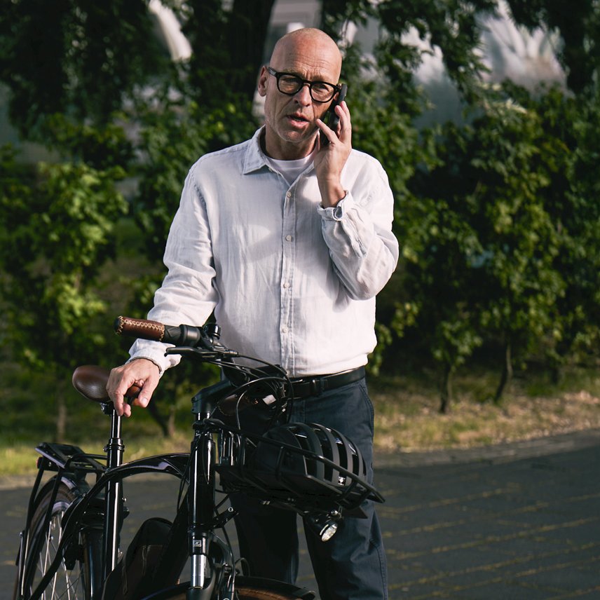 Ein Mann steht neben einem Fahrrad am Straßenrand und telefoniert mit seinem Smartphone. Im Hintergrund sind Bäume und eine ruhige Straße zu sehen.