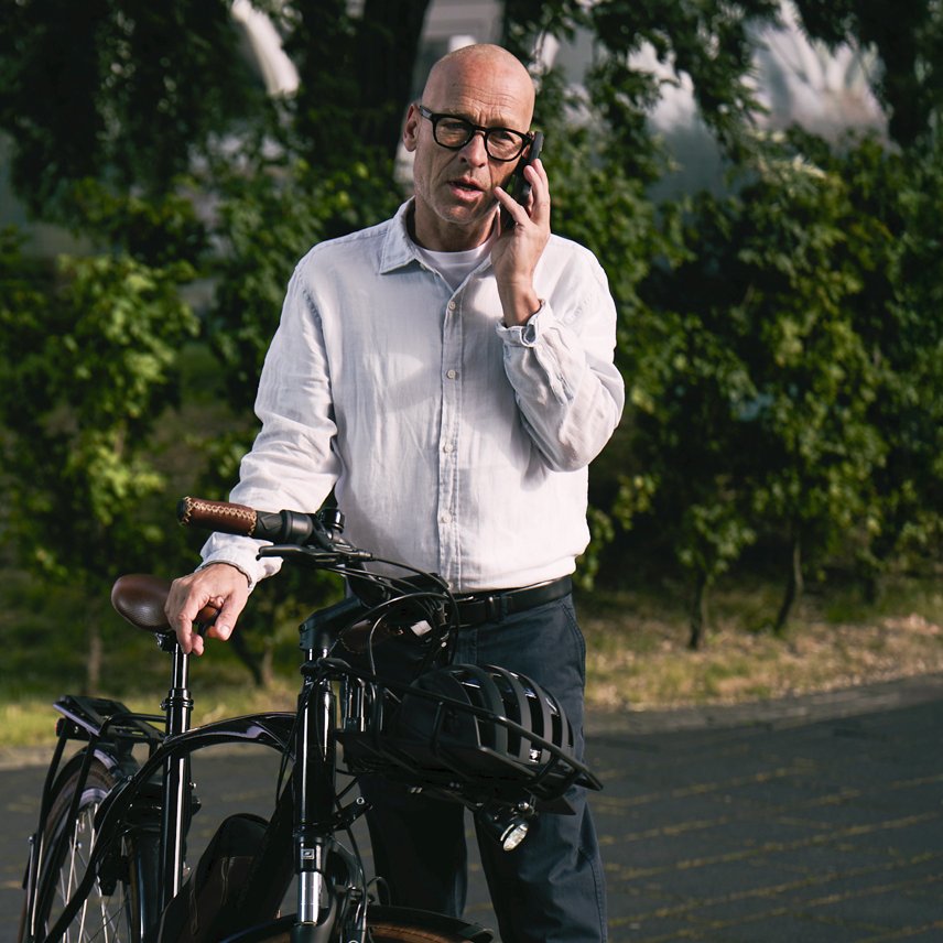 Ein Mann steht neben einem Fahrrad am Straßenrand und telefoniert mit seinem Smartphone. Im Hintergrund sind Bäume und eine ruhige Straße zu sehen.