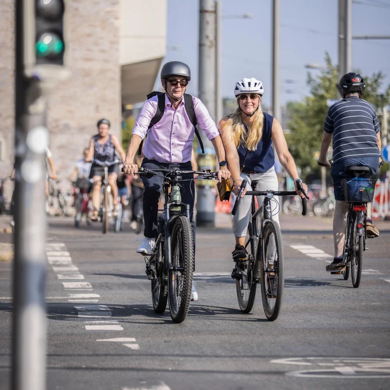 Mehrere Personen fahren mit Fahrrädern auf einer Straße in der Stadt. Zwei Radfahrende im Vordergrund tragen Helme und fahren nebeneinander.