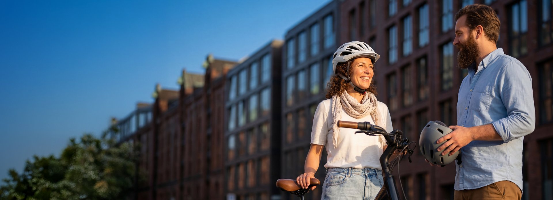 Eine Frau mit Fahrradhelm steht neben ihrem Fahrrad und spricht mit einem Mann, der einen Helm in der Hand hält. Im Hintergrund sind moderne Gebäude zu sehen.