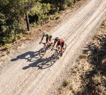 Zwei Radfahrende fahren auf einer hellen Schotterstraße durch eine bewaldete Landschaft, aufgenommen aus der Vogelperspektive.