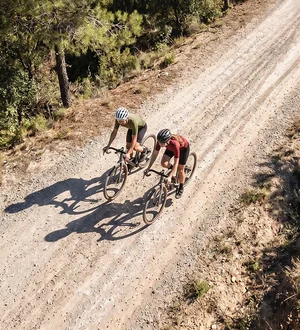 Zwei Radfahrende fahren auf einer hellen Schotterstraße durch eine bewaldete Landschaft, aufgenommen aus der Vogelperspektive.
