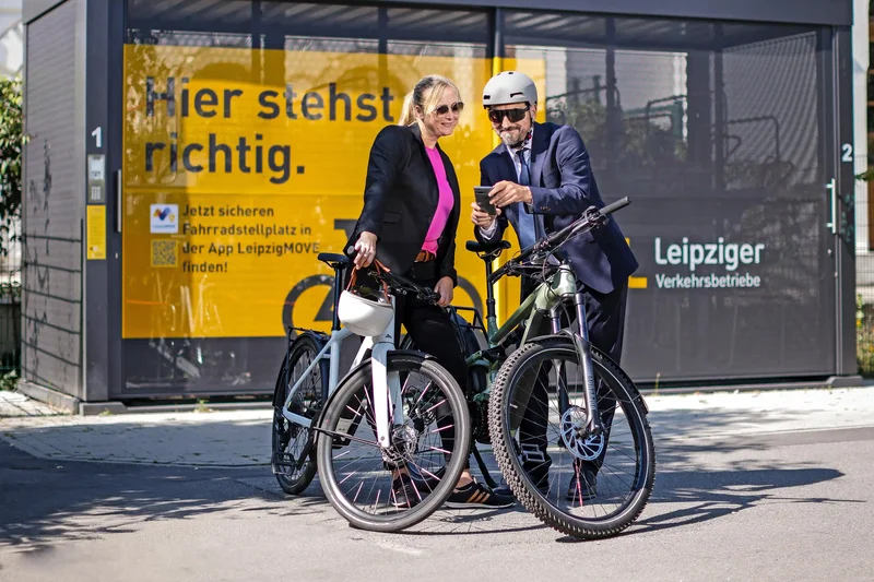 Zwei Personen stehen mit Fahrrädern vor einer Fahrradstation und schauen gemeinsam auf ein Smartphone. Im Hintergrund ist ein Gebäude der Leipziger Verkehrsbetriebe zu sehen.