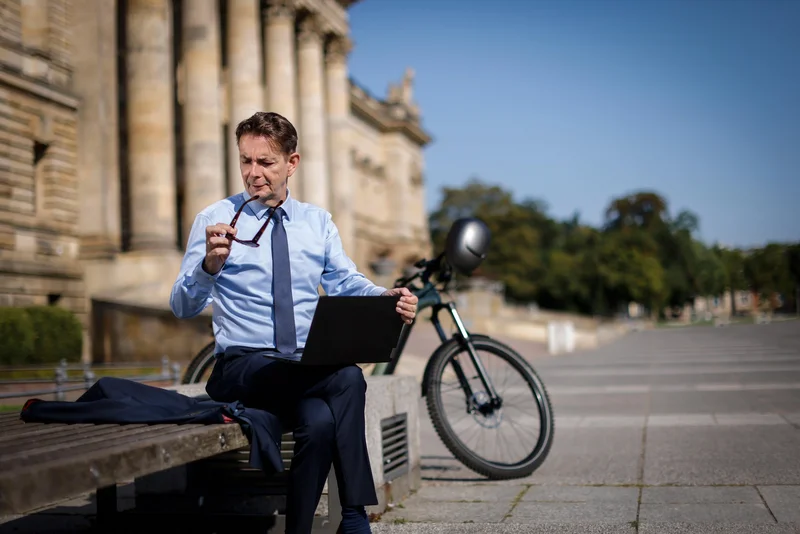 Ein Mann im Business-Outfit sitzt mit einem Laptop auf einer Bank im Freien. Neben ihm steht ein Fahrrad vor einem historischen Gebäude.