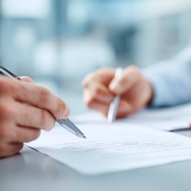 Ein Vertrag liegt auf dem Tisch, Hände zeigen mit Stiften auf den Vertrag.
