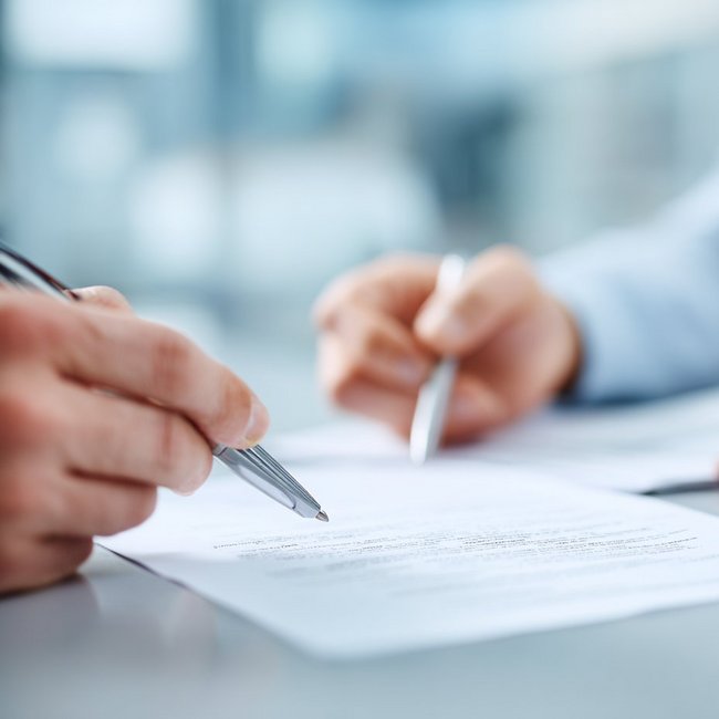 Ein Vertrag liegt auf dem Tisch, Hände zeigen mit Stiften auf den Vertrag.
