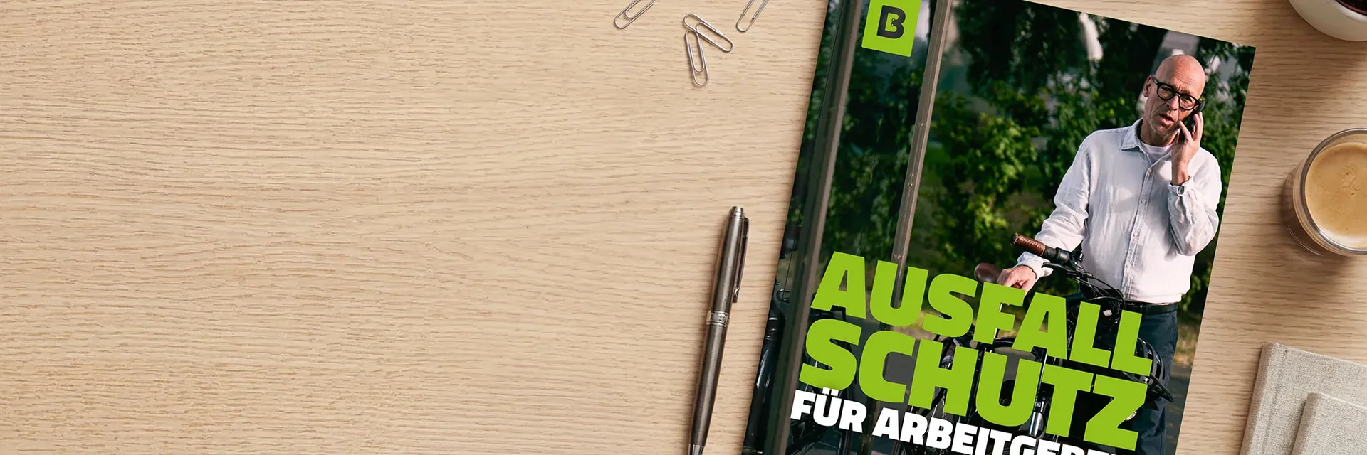 Auf einem Holztisch liegt ein Whitepaper mit der Aufschrift „Ausfallschutz“ und einem Foto eines Mannes mit Fahrrad; daneben sind ein Stift, Büroklammern und eine Kaffeetasse zu sehen.