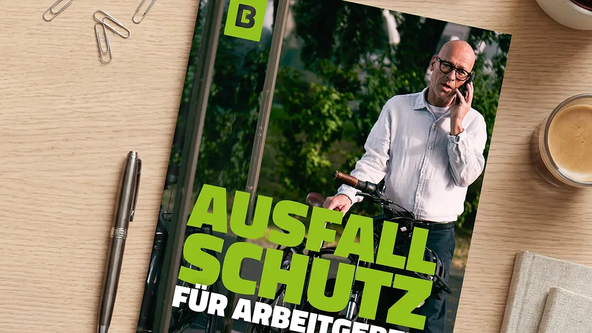 Auf einem Holztisch liegt ein Whitepaper mit der Aufschrift „Ausfallschutz“ und einem Foto eines Mannes mit Fahrrad; daneben sind ein Stift, Büroklammern und eine Kaffeetasse zu sehen.