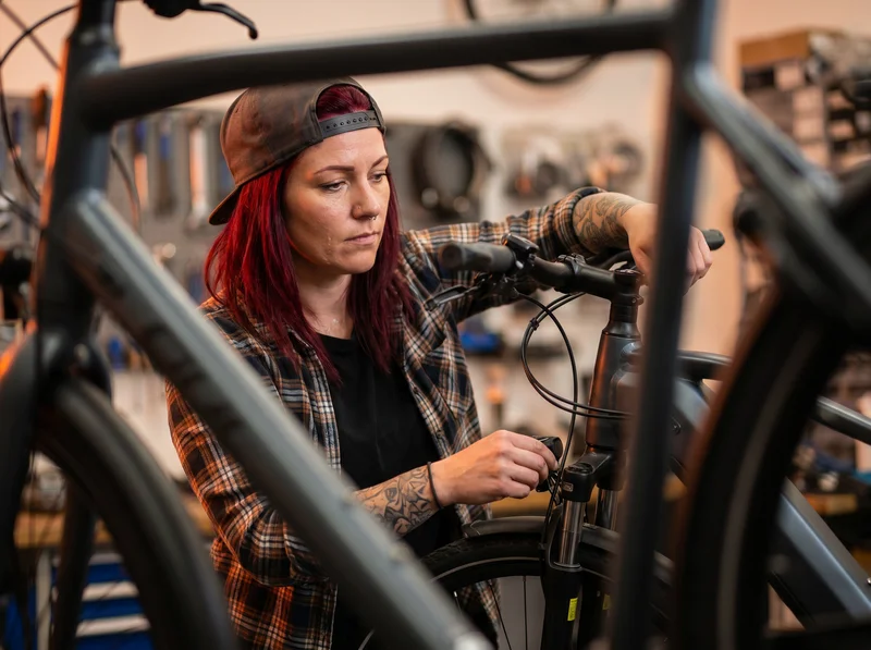Fahrradmechanikerin in einer Werkstatt justiert den Lenker eines Fahrrads; Werkzeuge und Ersatzteile sind unscharf im Hintergrund zu sehen.
