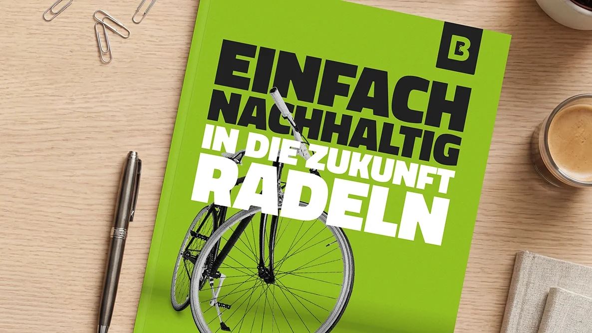 Auf einem Holztisch liegt ein grünes Whitepaper mit dem Titel „Einfach nachhaltig in die Zukunft radeln“ und einer Fahrrad-Abbildung; daneben sind ein Stift, Büroklammern und eine Kaffeetasse zu sehen.