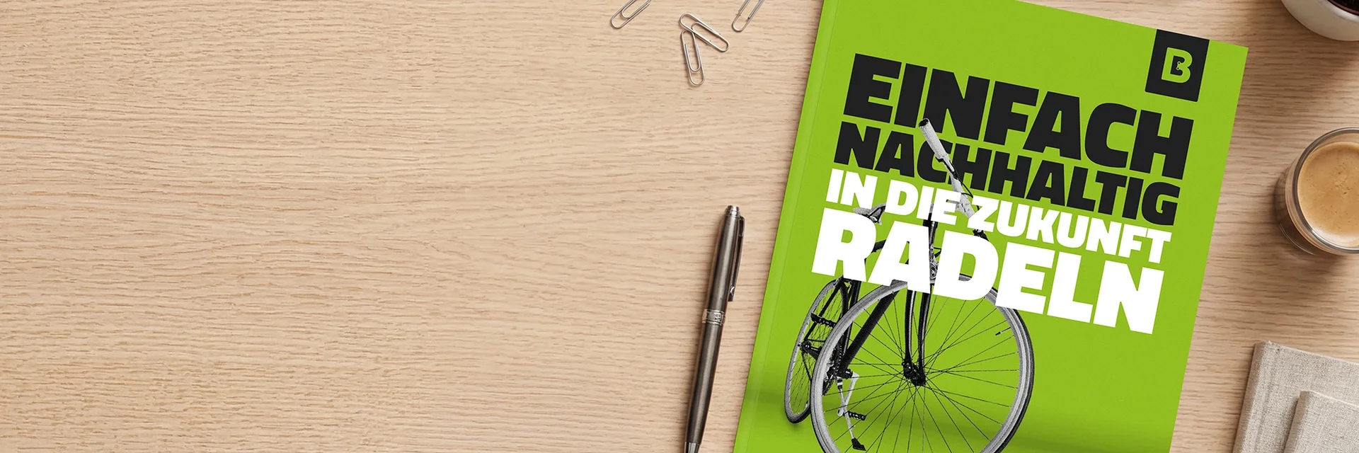 Auf einem Holztisch liegt ein grünes Whitepaper mit dem Titel „Einfach nachhaltig in die Zukunft radeln“ und einer Fahrrad-Abbildung; daneben sind ein Stift, Büroklammern und eine Kaffeetasse zu sehen.