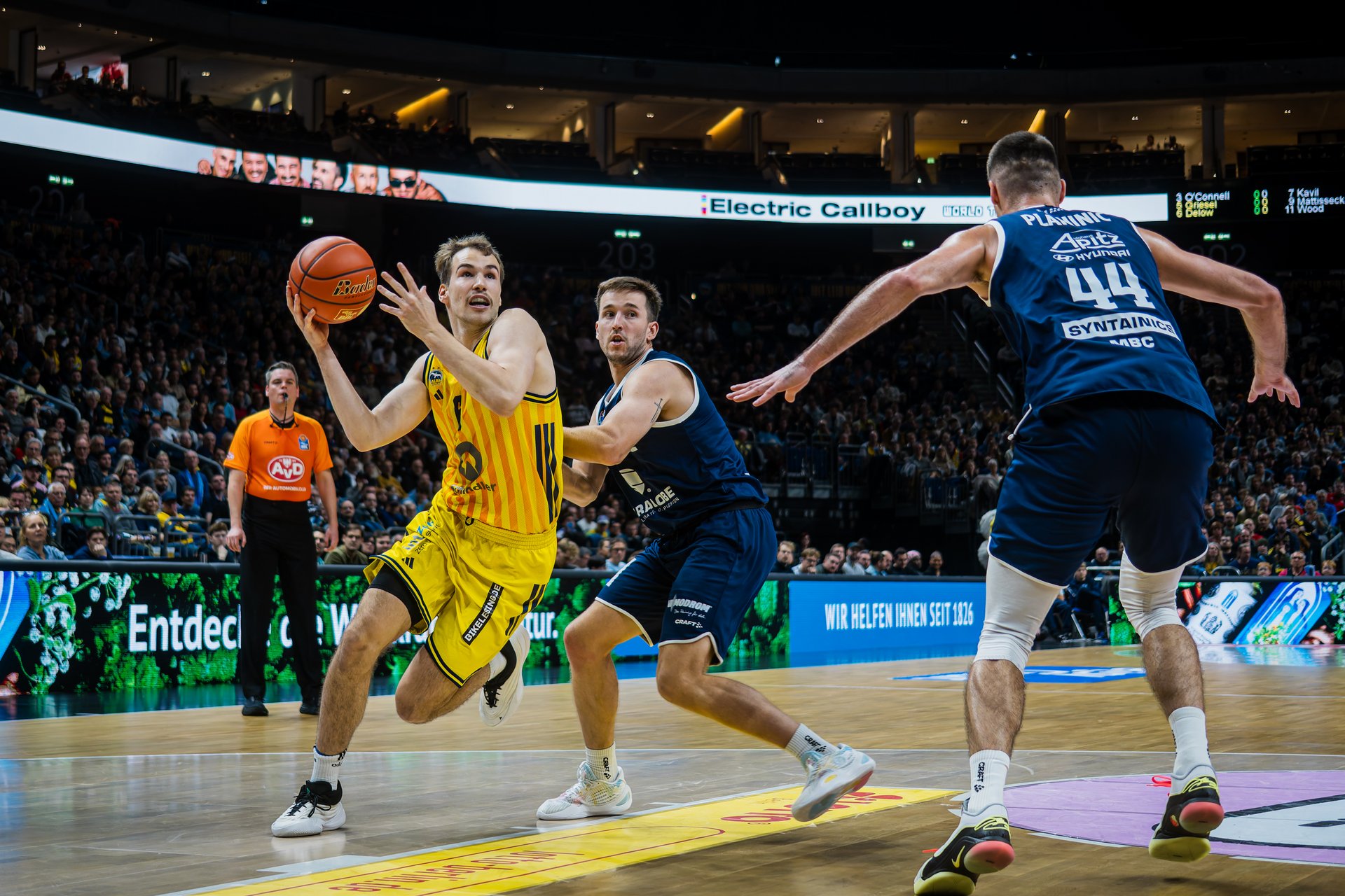 Basketballspiel in einer großen Halle: Ein Spieler im gelben Trikot springt mit dem Ball zum Korb, während zwei Gegenspieler in dunklen Trikots ihn verteidigen. Im Hintergrund sind Schiedsrichter, Spielfeldwerbung und zahlreiche Zuschauer zu sehen.