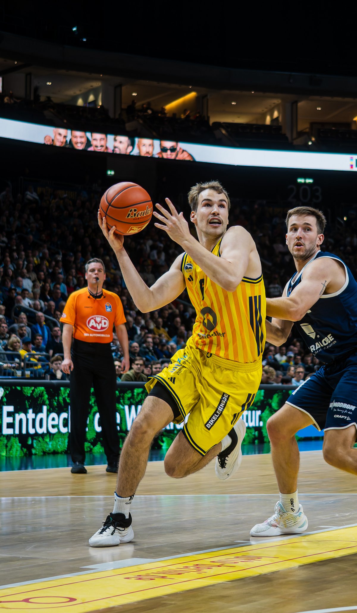 Basketballspiel in einer großen Halle: Ein Spieler im gelben Trikot springt mit dem Ball zum Korb, während zwei Gegenspieler in dunklen Trikots ihn verteidigen. Im Hintergrund sind Schiedsrichter, Spielfeldwerbung und zahlreiche Zuschauer zu sehen.