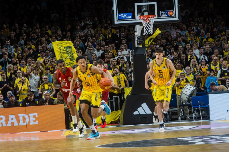Basketballspiel in einer voll besetzten Halle: Ein Spieler im gelben Trikot dribbelt den Ball nach vorne, verfolgt von Gegenspielern in roten Trikots. Im Hintergrund sind Korb, Spielfeldrand und zahlreiche Zuschauer zu sehen.
