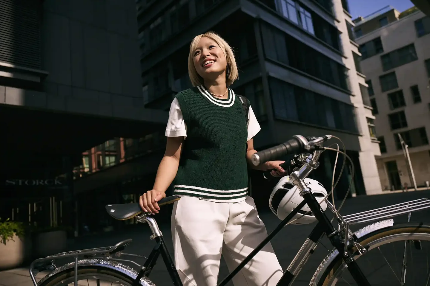 Junge Frau steht in der Stadt neben einem Fahrrad, hält den Lenker und blickt lächelnd nach oben; im Hintergrund sind moderne Gebäude zu sehen.