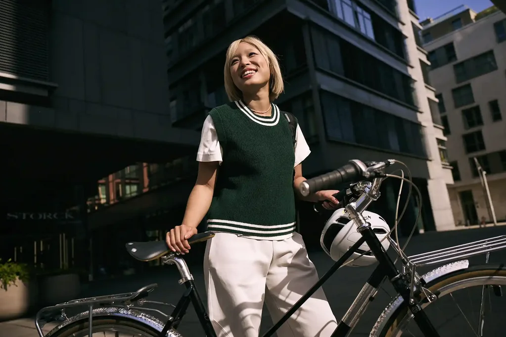 Junge Frau steht in der Stadt neben einem Fahrrad, hält den Lenker und blickt lächelnd nach oben; im Hintergrund sind moderne Gebäude zu sehen.