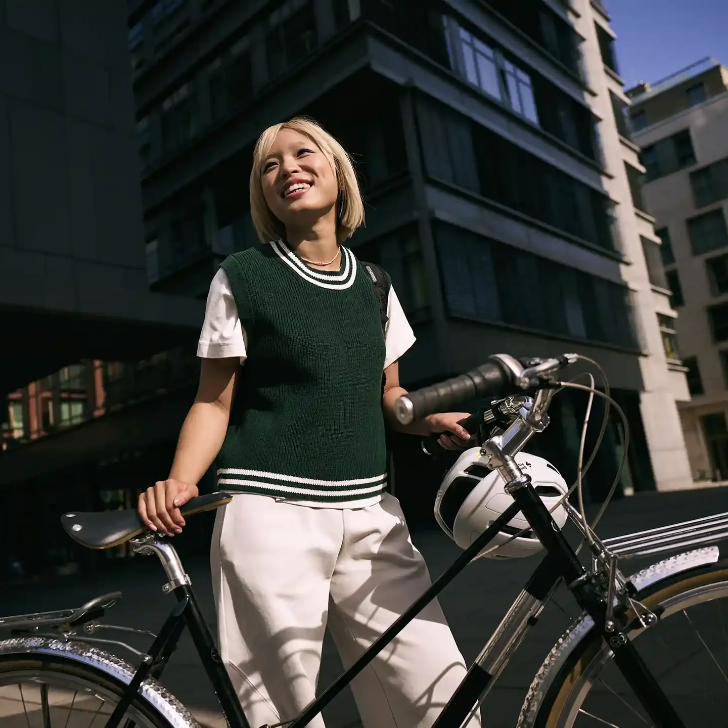 Junge Frau steht in der Stadt neben einem Fahrrad, hält den Lenker und blickt lächelnd nach oben; im Hintergrund sind moderne Gebäude zu sehen.