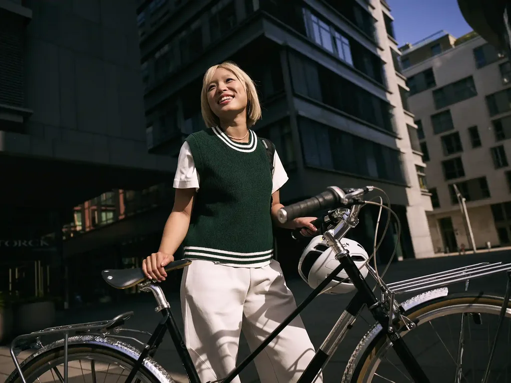 Junge Frau steht in der Stadt neben einem Fahrrad, hält den Lenker und blickt lächelnd nach oben; im Hintergrund sind moderne Gebäude zu sehen.